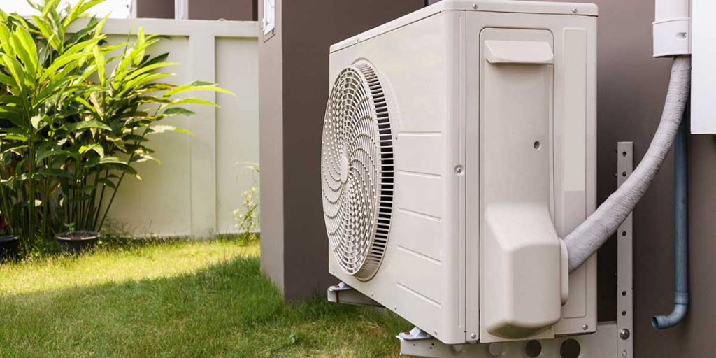 A modern outdoor air conditioner compressor unit is installed on a bracket next to a residential home's grassy backyard.