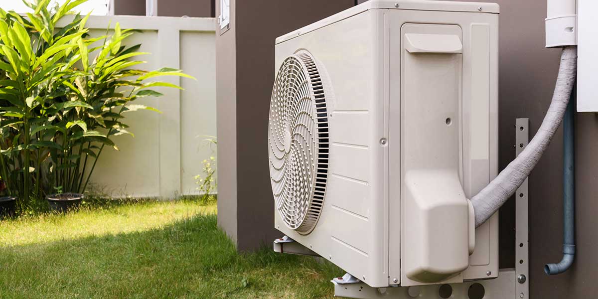 A modern outdoor air conditioner compressor unit is installed on a bracket next to a residential home's grassy backyard.