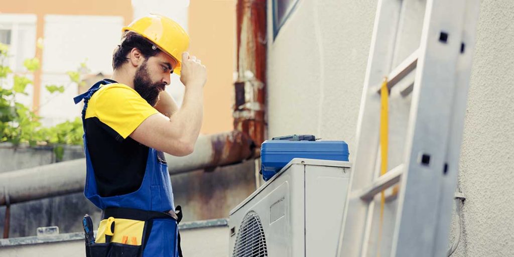 An HVAC technician adjusts his hard hat while inspecting an outdoor AC unit next to a ladder in San Jose.